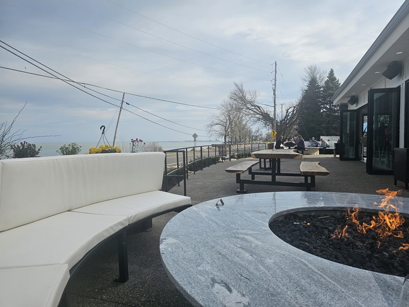 Patio table with a firepit in the center, with couch seating to the left and view of the lake in the background