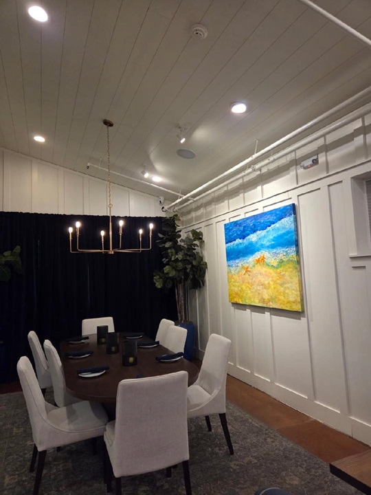 Large table in an interior room, surrounded by white chairs, with a large painting on the wall
